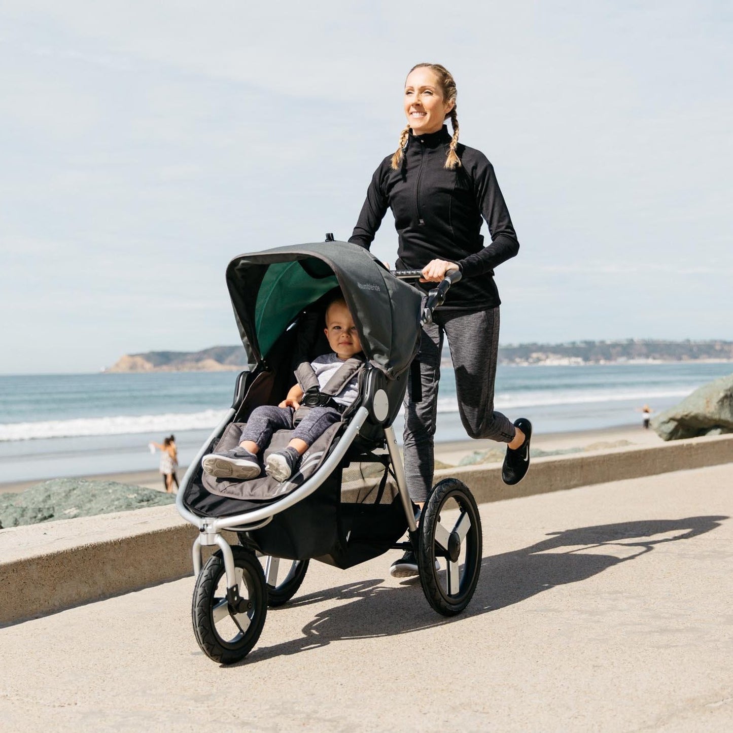 Young mother running along a beachfront while pushing a black and grey stroller. Inside the stroller is a child that is slightly smiling.